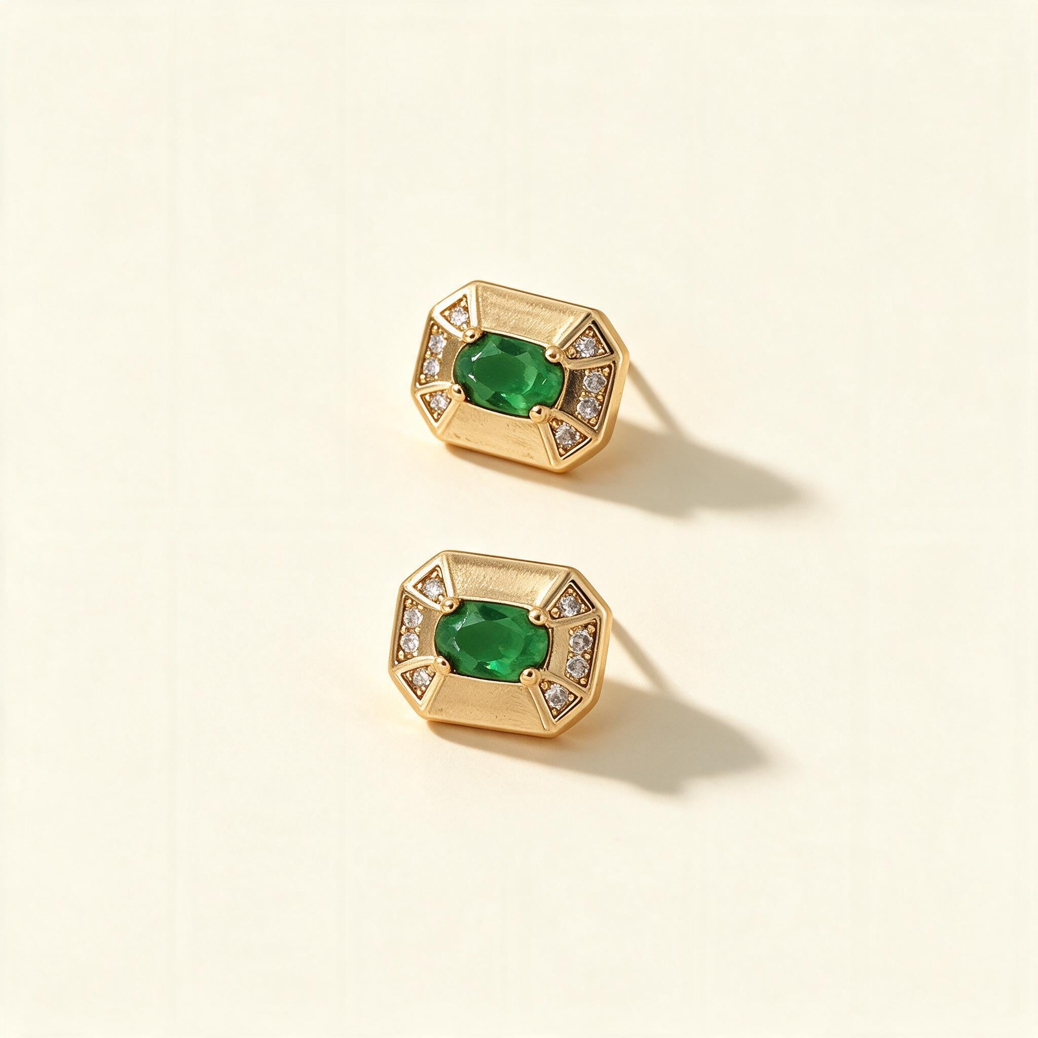Gold-tone octagonal stud earrings with emerald green oval stone and cubic zirconia corners on ivory background