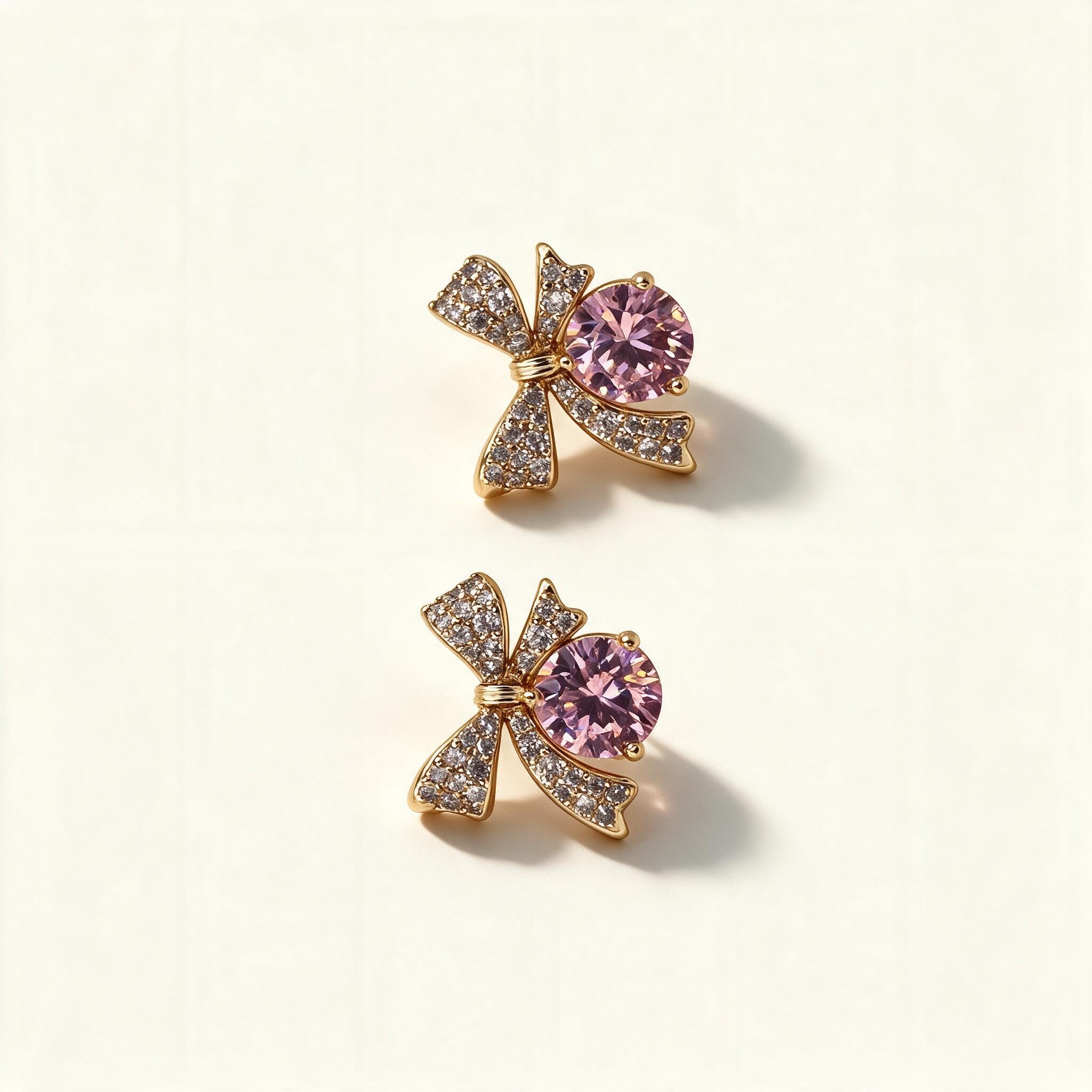 Gold-tone pave bow stud earrings with a large round pink cubic zirconia on ivory background