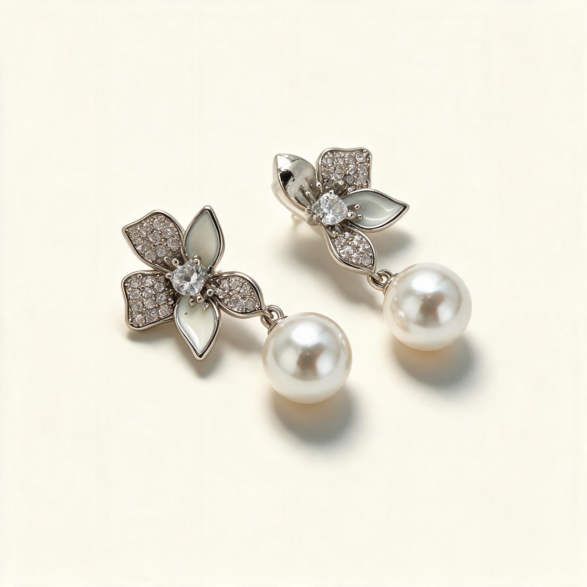 Silver-tone flower earrings with cubic zirconia and white enamel petals and a hanging shell pearl on ivory background