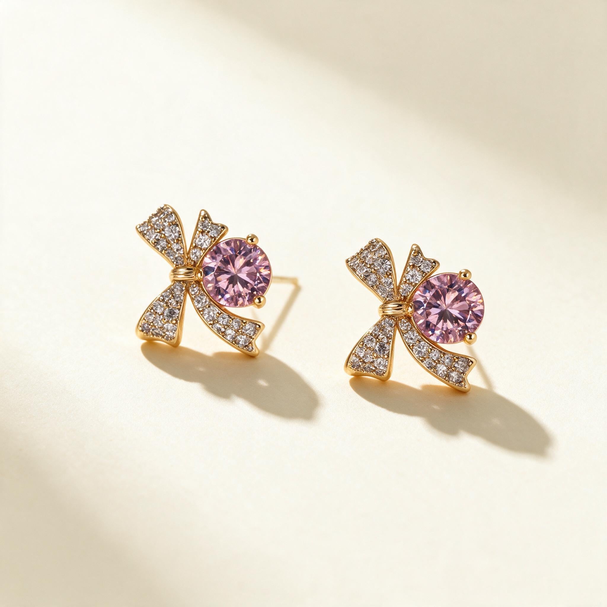 Gold-tone pave bow stud earrings with a large round pink cubic zirconia on ivory background