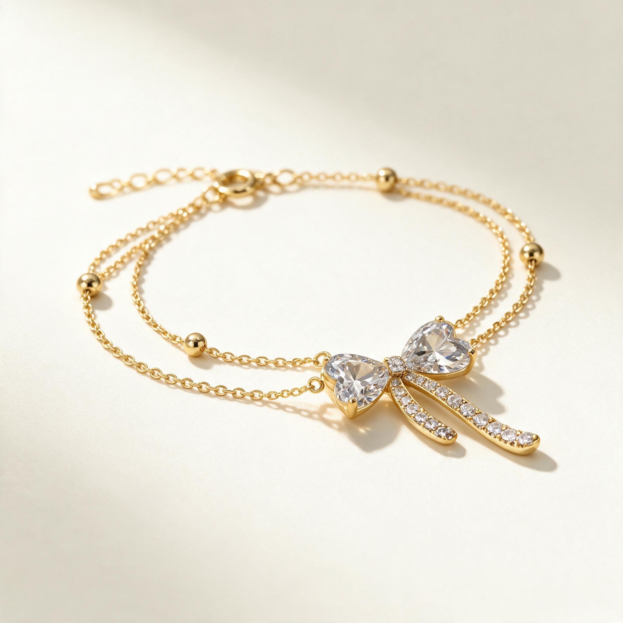 Gold-tone double chain bracelet with heart cubic zirconia bow charm and bead stations on ivory background