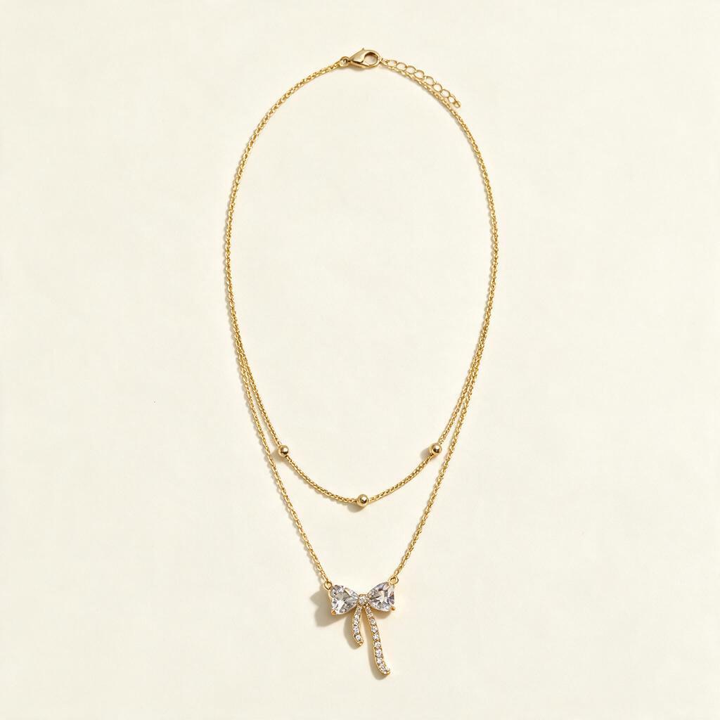 Gold-tone double chain necklace with a clear crystal bow and pavé ribbon tails on ivory background